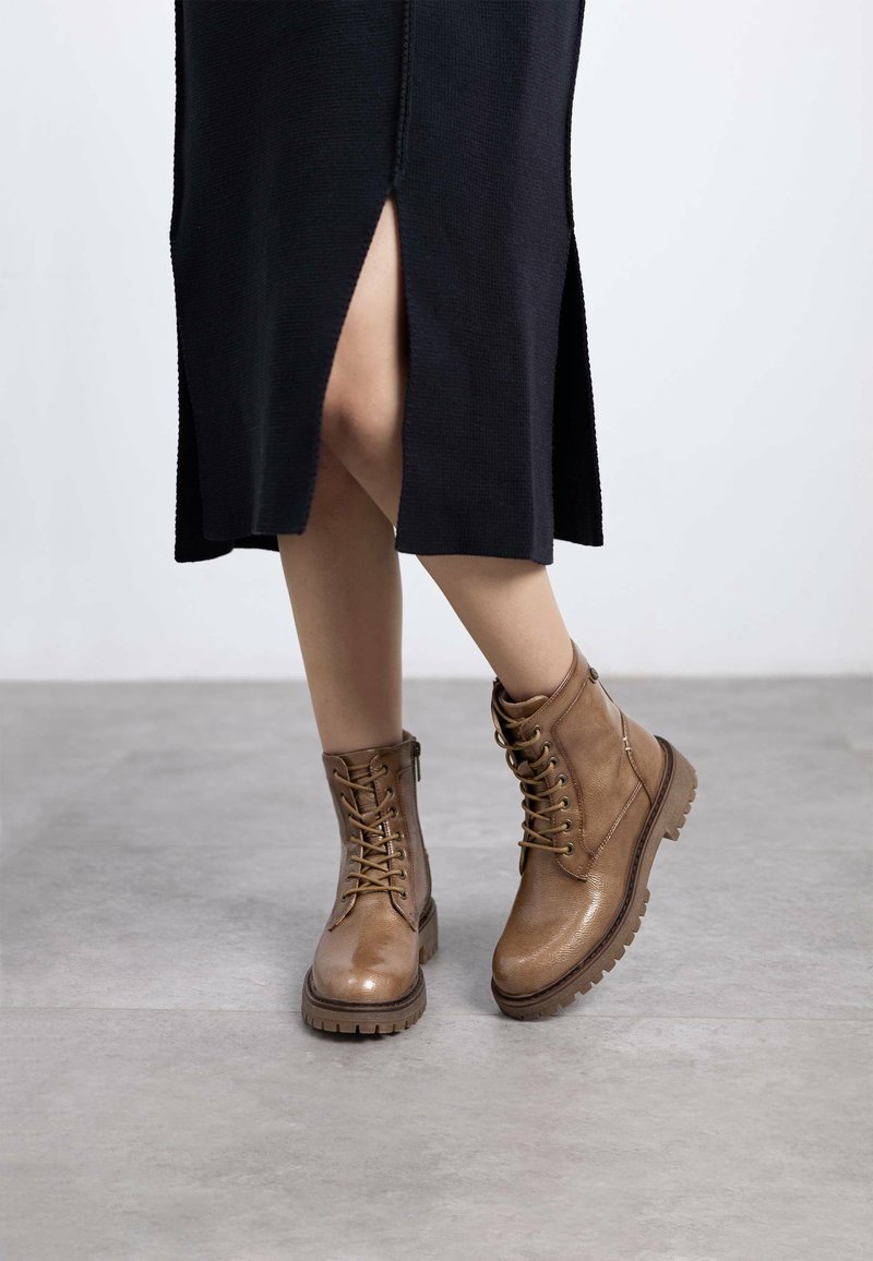 Botas de tobillo de cuero marrón con cordones, acabado texturizado, puntera redondeada y suela robusta, combinadas con un vestido negro que presenta aberturas laterales.