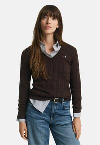 Brauner, kabelgestrickter Pullover mit V-Ausschnitt, getragen über einem hellblauen gestreiften Hemd. Kombiniert mit blauen Jeans und einem schwarzen Gürtel.