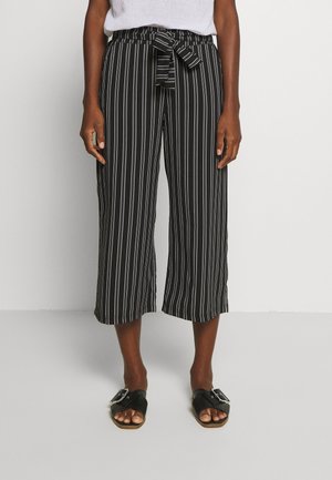 Person trägt schwarze, gekürzte Hose mit weißen vertikalen Streifen und einem gebundenen Gürtel, kombiniert mit schwarzen Slip-On-Sandalen.