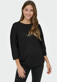 Mujer con cabello largo y ondulado sonriendo, vistiendo una blusa negra de corte holgado con mangas de tres cuartos y jeans negros desgastados.