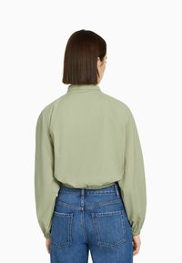 Light green, textured jacket with a high collar and gathered cuffs, paired with blue denim jeans, showing back view and seam details.