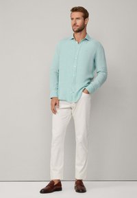 Mintgroene button-up shirt van lichtgewicht stof, gecombineerd met witte slim-fit broek en bruine instappers, handen in de zakken, neutrale achtergrond.