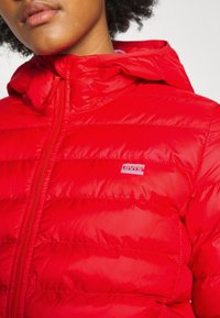 Chaqueta acolchada roja con acabado brillante, que incluye capucha, cierre con cremallera y costuras horizontales. Pequeño logo blanco de Levi's en el pecho.