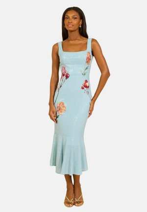 Kvinde iført en lyseblå ærmeløs kjole med blomsterbroderi og udvidet skørt, kombineret med beige sandaler med remme.