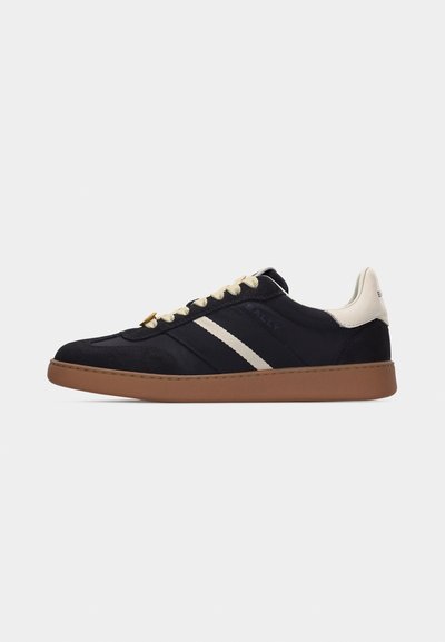 Baskets basses noires avec lacets beige, languette blanche au talon, rayures blanches sur les côtés et semelle en caoutchouc marron sur un fond clair uni.