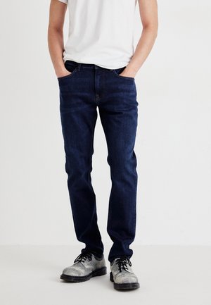 Mężczyzna ubrany w ciemnoniebieskie dżinsy i szare buty sznurowane, z rękami w kieszeniach, stoi na tle jednolitego białego tła.