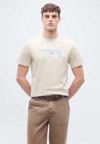 Νεαρός άνδρας που φοράει μπεζ μπλουζάκι Calvin Klein με μπλε κείμενο και χακί παντελόνι με καφέ ζώνη, στέκεται μπροστά σε απλό φόντο.