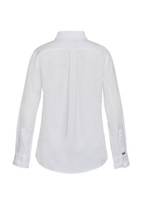 Chemise blanche à manches longues avec un pli central au dos, des poignets boutonnés et une petite étiquette rouge, blanche et bleue sur le poignet gauche.