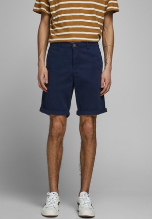 Marineblaue Baumwollshorts mit aufgerolltem Saum, ausgestattet mit einem Knopf- und Reißverschluss vorne. Kombiniert mit einem braun-weiß gestreiften Hemd.