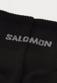 Calcetín negro con textura acanalada; presenta la palabra "SALOMON" en letras grises de punto en la parte superior.