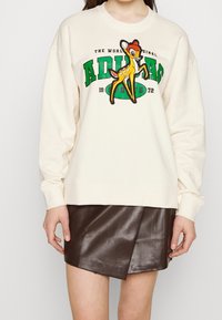 Krämfärgad sweatshirt med rund halsringning, prydd med en broderad tecknad hjort i gult och rött, samt grön text. Matchas med en kjol i brunt läder.