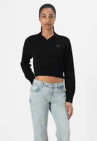 Black cropped sweater with a V-neckline, ribbed cuffs and hem, featuring a small black heart accent on the chest. Worn with light blue jeans.