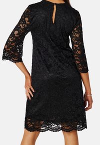 Robe noire en dentelle avec manches trois-quarts, dos en forme de goutte et ourlet festonné. Présente un motif floral et une superposition texturée.