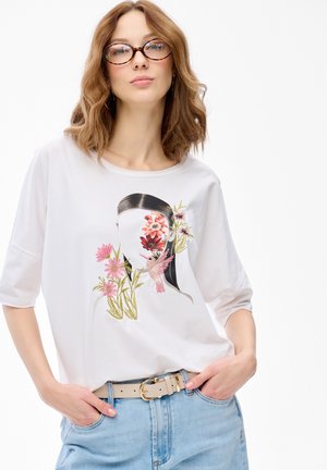 Kobieta o falowanych, jasnobrązowych włosach i okularach, ubrana w białą koszulę z wzorem kwiatów i ptaków na twarzy, jasnoniebieskie dżinsy oraz beżowy pasek.