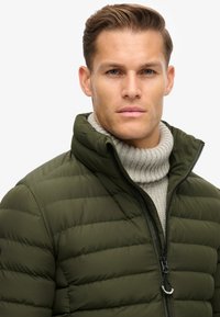 Olivengrøn pufferjakke med høj krave, vandret syninger og lynlås foran. Bæres over en lysegrå ribbet turtleneck sweater.