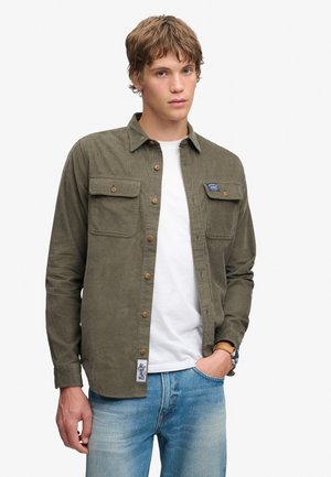 Junger Mann trägt ein olivgrünes Hemd mit Brusttaschen, ein weißes T-Shirt, hellblaue Jeans und eine Armbanduhr, posiert mit einer Hand in der Hosentasche.