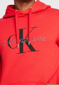 Punainen puuvillaneule huppareilla, jossa on säädettävä huppu. Esittää suurta mustaa "CK"-logoa ja "CALVIN KLEIN JEANS" -tekstiä hopeakirjailulla.
