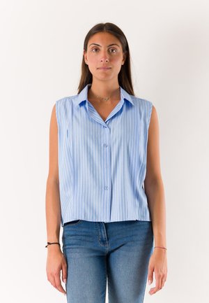 Donna in piedi di fronte, indossando una camicia senza maniche a righe bianche e blu con bottoni e jeans blu su uno sfondo semplice.