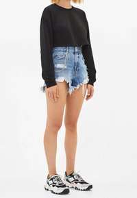 Mujer que lleva una sudadera negra corta, pantalones cortos de denim rasgados de cintura alta con detalle de cadena, y zapatillas coloridas y gruesas, de pie frente a un fondo blanco.