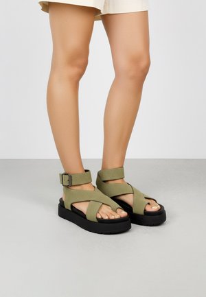 Piernas con pantalones cortos beige y sandalias de plataforma verdes con anchos tirantes cruzados y hebillas en el tobillo sobre un suelo gris.