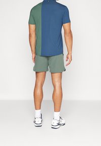 Camisa polo de manga corta en azul y verde, combinada con pantalones cortos verdes. La tela parece suave con un ajuste relajado. Zapatillas deportivas blancas visibles.