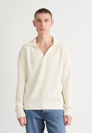Suéter de punto color crema con cuello en V y cuello ancho; presenta un patrón texturizado y puños acanalados. Combinado con jeans de mezclilla azul.