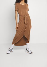Robe midi marron à manches courtes avec une ceinture à nouer à la taille et un avant drapé. Portée avec des chaussettes blanches et des chaussures de sport. Texture lisse.