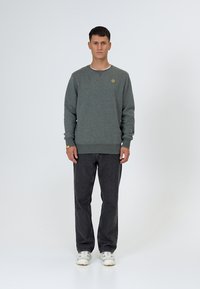 Sweat-shirt en coton gris avec un col rond et des poignets côtelés, orné d'un petit logo jaune. Porté avec un jean foncé et des baskets blanches.