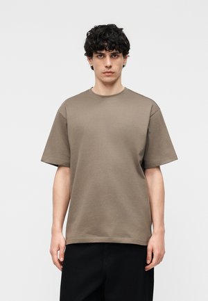 Jeune homme aux cheveux bouclés et foncés portant un t-shirt marron uni à manches courtes et un pantalon noir, debout devant un fond clair uni.
