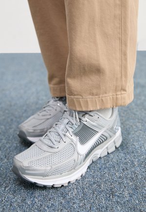 Scarpe da corsa Nike grigie con logo swoosh bianco indossate con pantaloni beige su pavimento moquettato blu.
