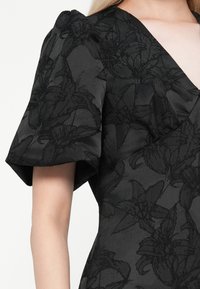 Čierne šaty s textúrovaným kvetinovým vzorom. Majú krátke pufované rukávy a V-výstrih. Hladká látka s jemným leskom.