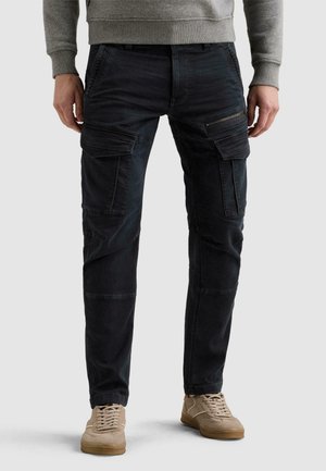 Homme portant un pantalon cargo noir avec plusieurs poches et des baskets beiges, debout devant un fond clair uni.