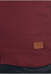 Bordo pleteni pulover s rebrastim rubom i kožnom oznakom s utisnutim tekstom blizu dna. Glatka tekstura.