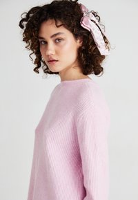 Roze geribd trui met een hoge halslijn en lange mouwen. Voorzien van een grote roze satijnen strik met versieringen aan de zijkant.