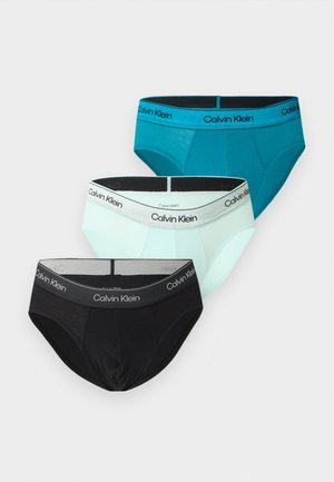 Męski model o ciemnej karnacji, ubrany w jasnoniebieskie majtki Calvin Klein, stoi boso z jedną ręką na biodrze na białym tle.