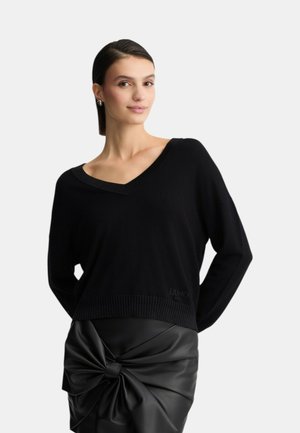 Maglione nero a V con orlo a coste, realizzato in morbido materiale. Presenta un logo vicino al fondo. Abbinato a una gonna in pelle nera.