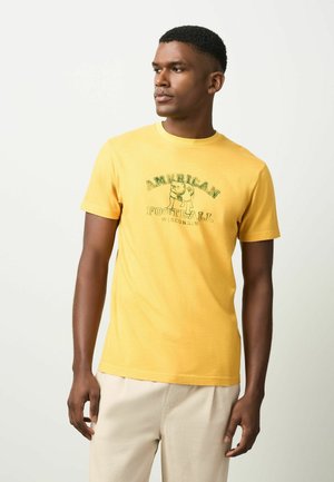 Man wearing a yellow T-shirt with text "American Football Wisconsin" and an illustrated bulldog, standing against a plain white background.
