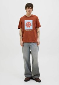 Brun bomulds t-shirt med et blåt firkantet grafik, der har et rødt sol-design. Paireret med løst siddende grå jeans og brune støvler.