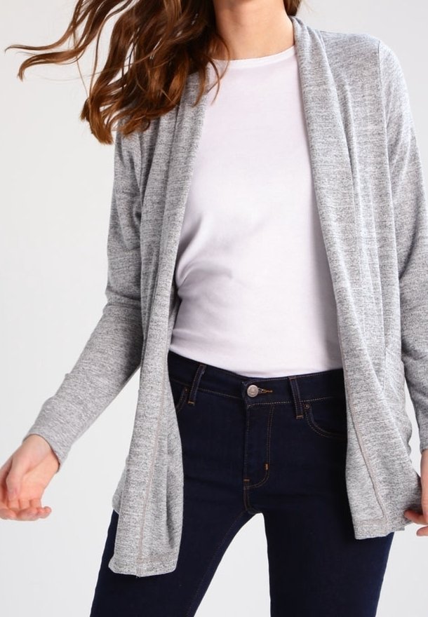 Femme portant un cardigan gris clair ouvert sur le devant, un t-shirt blanc à col rond, et un jean bleu foncé, visible du cou jusqu'aux hanches.