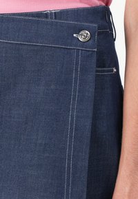 Mørk denimskjørt med knappelukking, har kontrasterende hvit søm og en sidelomme. Materialet er teksturert og slitesterkt.