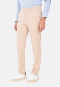 Pantaloni eleganti beige con vestibilità slim, dotati di passanti per cintura, tasche laterali e una trama di tessuto liscia, abbinati a sneaker bianche.