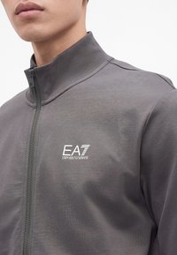 Млад човек, носещ сива яке с цип и висока яка и бяло лого EA7 Emporio Armani на гърдите.