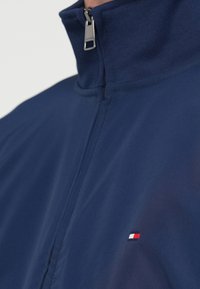 Σκούρα μπλε μπουφάν με φερμουάρ, που διαθέτει ψηλό γιακά, λεία υφή και μικρό λογότυπο σε κόκκινο, άσπρο και μπλε στην αριστερή πλευρά του στήθους.