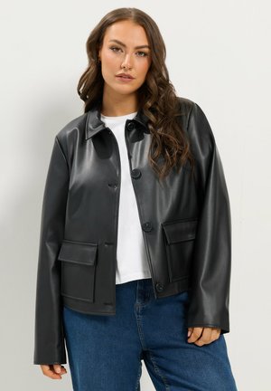 Donna con lunghi capelli ondulati che indossa una giacca di pelle nera sopra una camicia bianca e jeans blu su uno sfondo chiaro e uniforme.