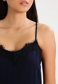 Navy satin linne med intrikata svarta spetsdetaljer längs halsringningen och delikata fransar vid kanterna. Spaghettiband.