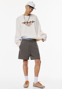 Cremfarbener Oversize-Pullover mit braunem Grafikdesign, graue Shorts, beige Sneakers mit orangefarbenen Schnürsenkeln, weiße Socken und eine weiße Mütze.