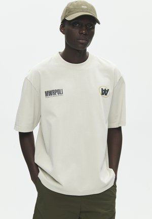 Young man wearing a beige cap and off-white T-shirt with black and yellow logos, hands in pockets, standing against a plain light background.