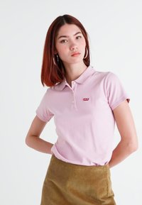 Polo de color rosa claro con mangas cortas, tapeta de dos botones y un pequeño logo rojo. Combinado con una falda de pana color caqui.