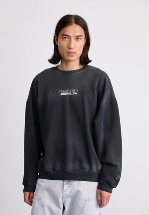 Dark gray sweatshirt with dropped shoulders, ribbed cuffs, and hem. Features white "CAVERTENE BERLIN" logo on the chest. Smooth texture.