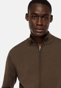 Brown zip-up sweater with a high collar and ribbed texture, featuring a silver zipper and a fitted design.